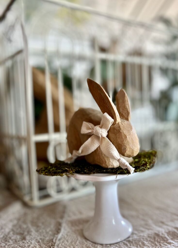 super closeup of a carved wood bunny with a bow around his neck on dried moss on a white cupcake stand with the decorative birdcage behind