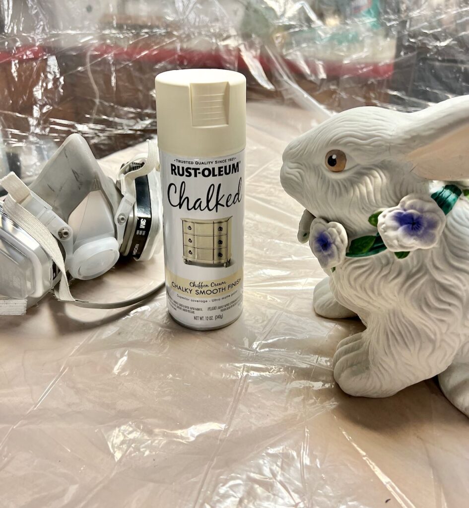 image of the original bunny statue with the bright colored flower necklace and scary painted eyes, and bottle of rust-oleum brand chalk spray paint and a respirator in front of a large plastic drop clothh