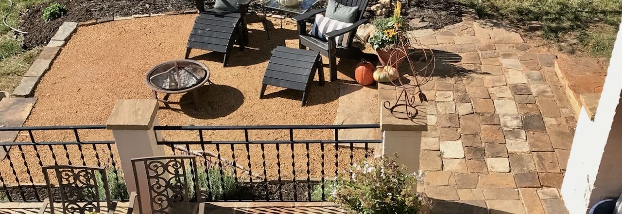 A Wide Title Shot of the new DG patio with hchairs and a firepit, the stone walkway next to it and the edge of the brick patio with iron railing and brick posts also seen