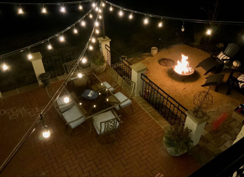 view from above on. the balcony looking down over a roaring fire in the fire pit on the new gravel patio and strings of lit outdoor Edison bulbs hanging over a red brick patio