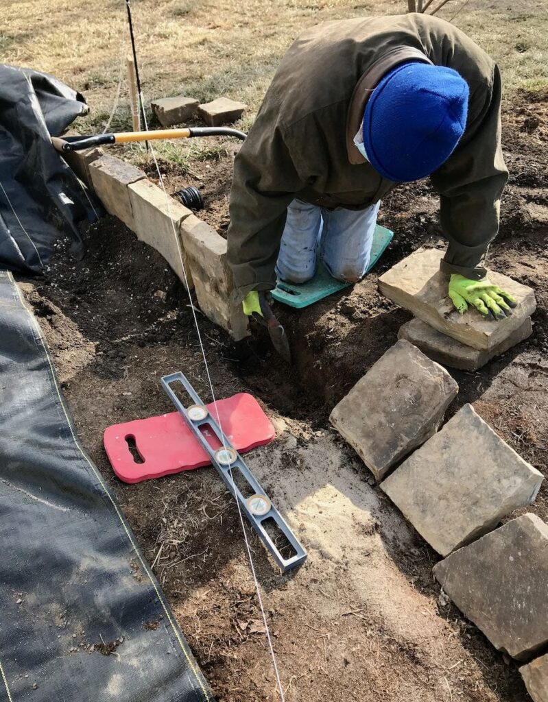 Man kneeling beside a narrow trench in the dirt placing each stone vertically to create a barrier between the DG Patio and the landscape berm