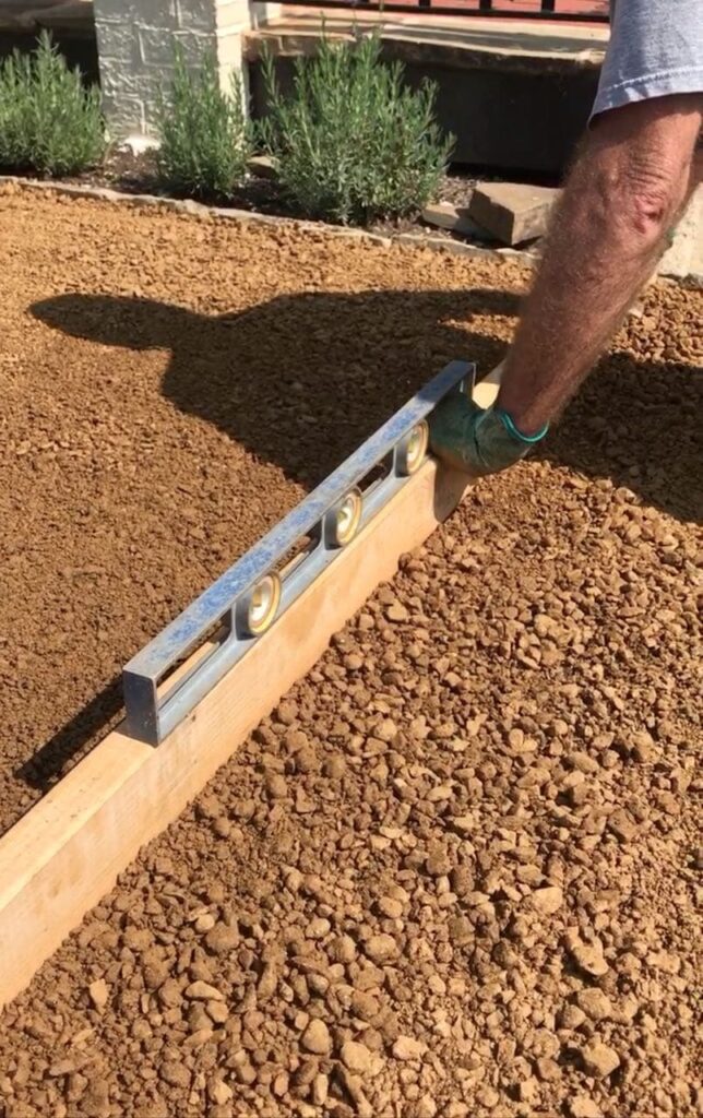 Man arm showing holding a level on a long 2x4 pushing it across the DG to level it final layer