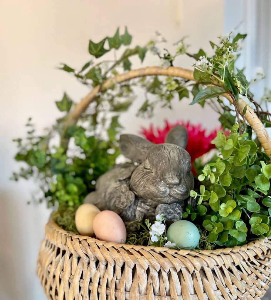 Sweet "concrete" bunny is sleeping on a bed of moss in a large basket with vines growing up the handle and three soft-colored eggs around
