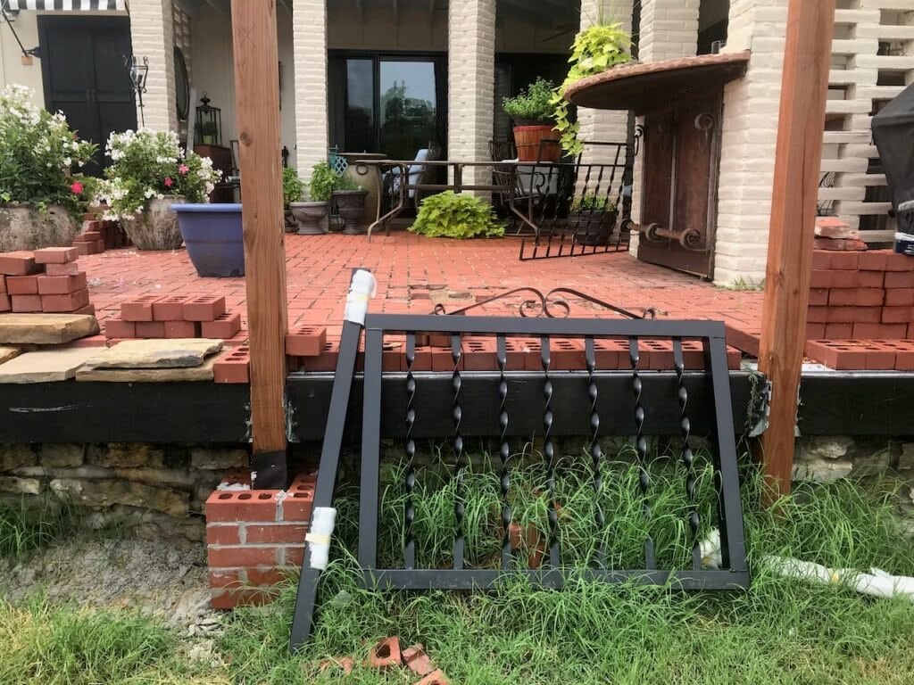 a block iron gate is leaning against the edge of an elevated red brick patio with a black facing board hung between two two posts