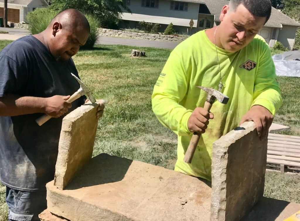 Two men using hammers to intentionally chip the edges of natural stone caps for the new brick columns