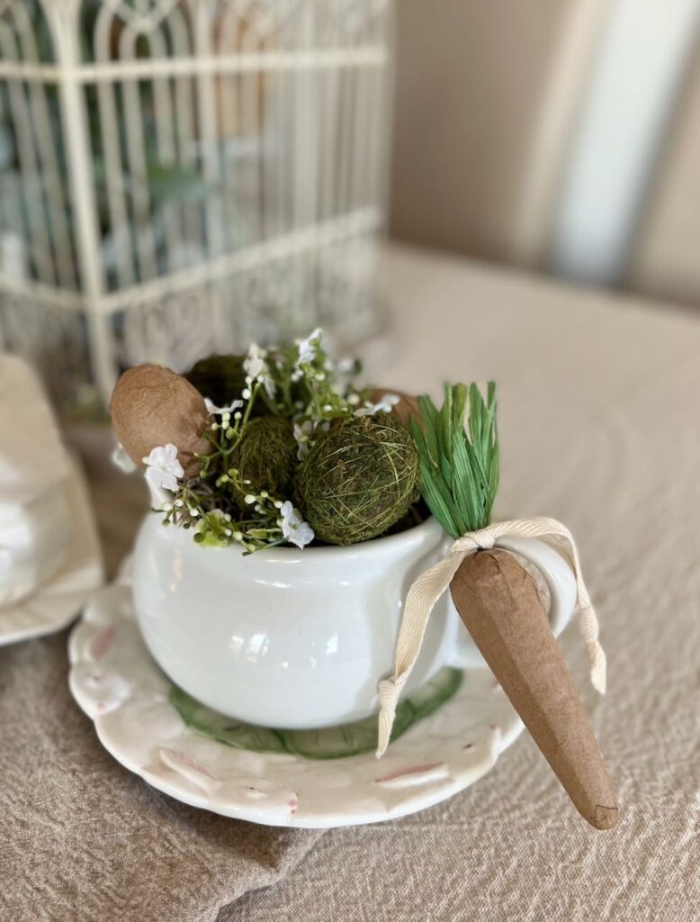 a white soup mug is on a bunny salad plate and filled with dried moss with paper mache eggs and moss eggs. The handle is accented with a paper mache carrot tied on it.