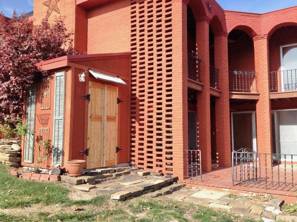 Before Picture of the back of the house five two-story brick arches supporting a balcony with iron railing wrapping the north and west sides of a brick patio below that has. the same short black iron. railing. This view shows the tool shed attached to the side of the house