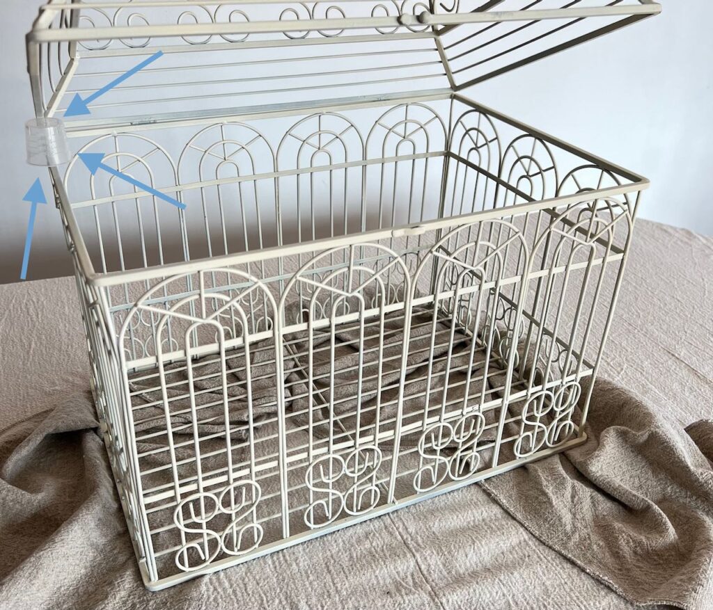 Birdcage shown with the lid propped open and three arrows pointing out the clear plastic medicine cup sitting. on. the side rail with the top propped on top