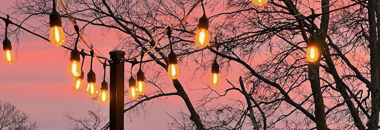 wide closeup view of dour strands of lit outdoor Edison bulbs suspended from a black metal pole with a dramatic pink and purple sky behind