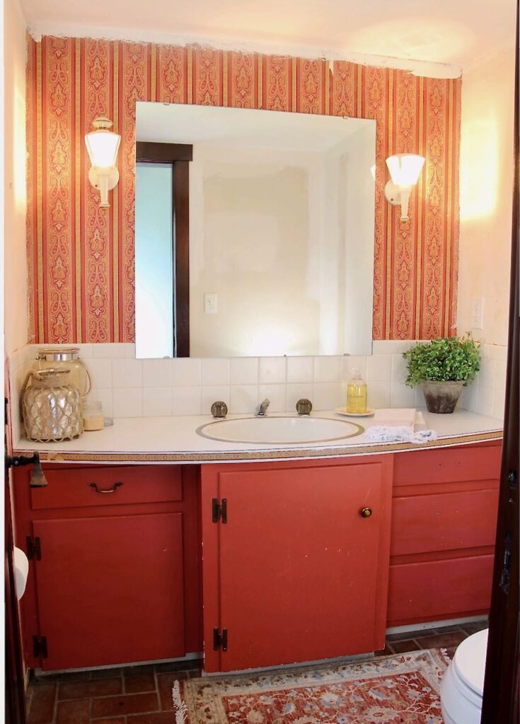 view of the original vanity and mirror with thee wallpaper cornice board reemoved and the toilet and door moved