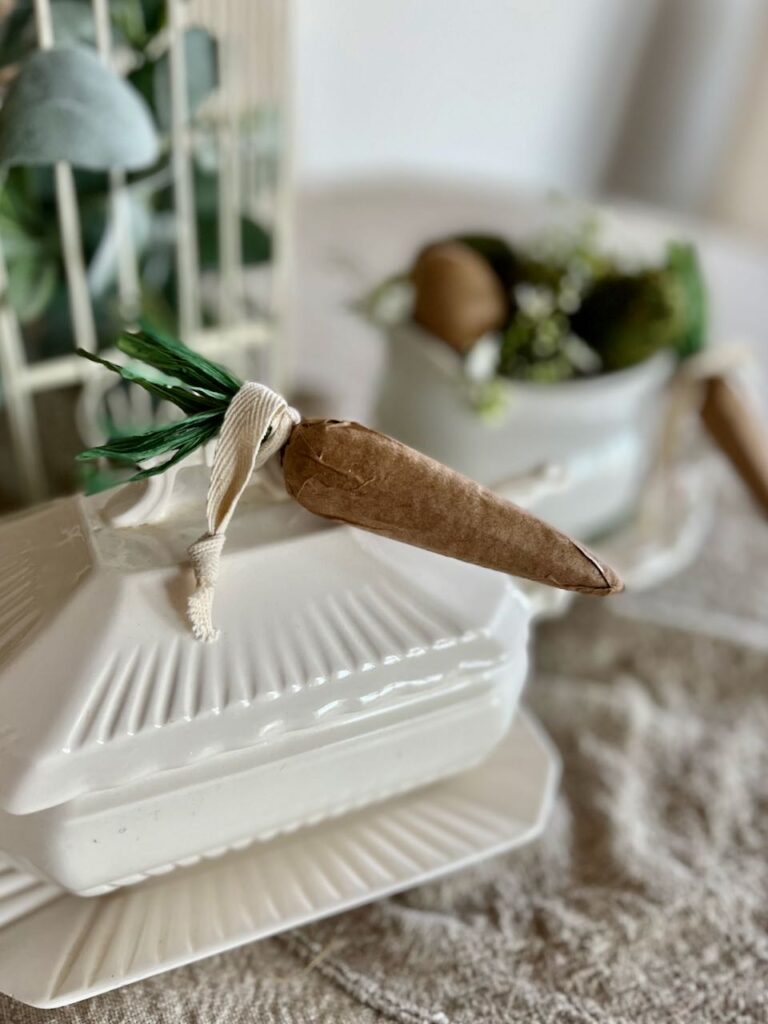 closeup of a paper mache carrot tied to the handle of the lid of a sauce boat with a soup mug decorated with eggs and moss in the backgroound