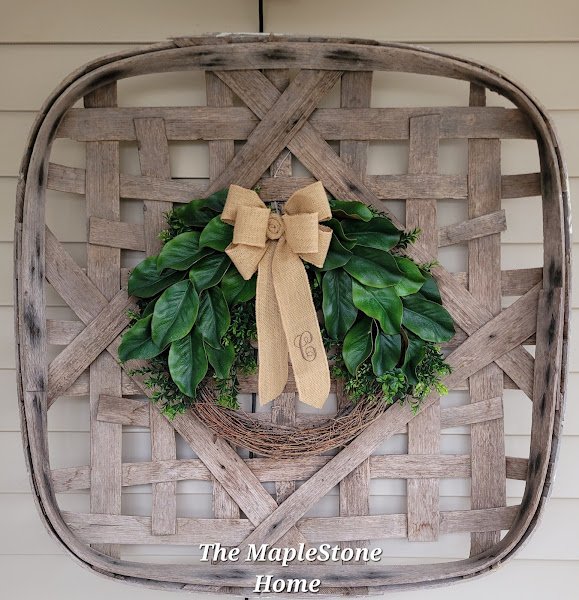 A grapevine wreath with magnolia leaves on top with a monogrammed burlap bow all mounted on a vintage tobacco basket