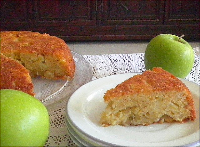 a slice of apple cake is on a plate with the rest of the cake in the background and two Granny Smith apples close by