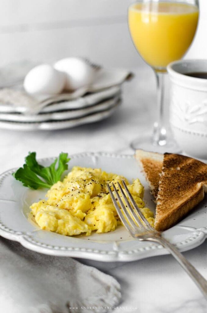 golden yellow scrambled eggs on a white plate with a fork and some toast with a glass of milk and orange juice in the background