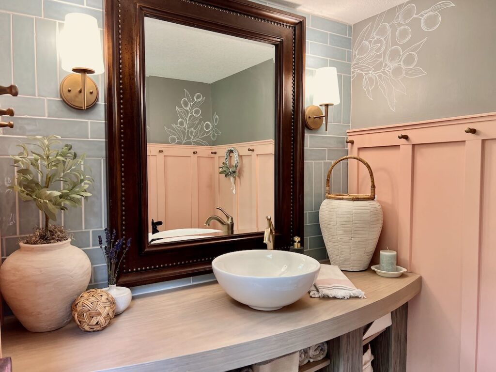 Image of the finished powder room remodel showing the vanity top with sink, faucet and decor pieces in front of a blue tile wall and a large framed mirror. Reflected in the mirror is the coral board and batten wall with gold pegs on on one peg is hanging a small wreath accented with dried lambs ear