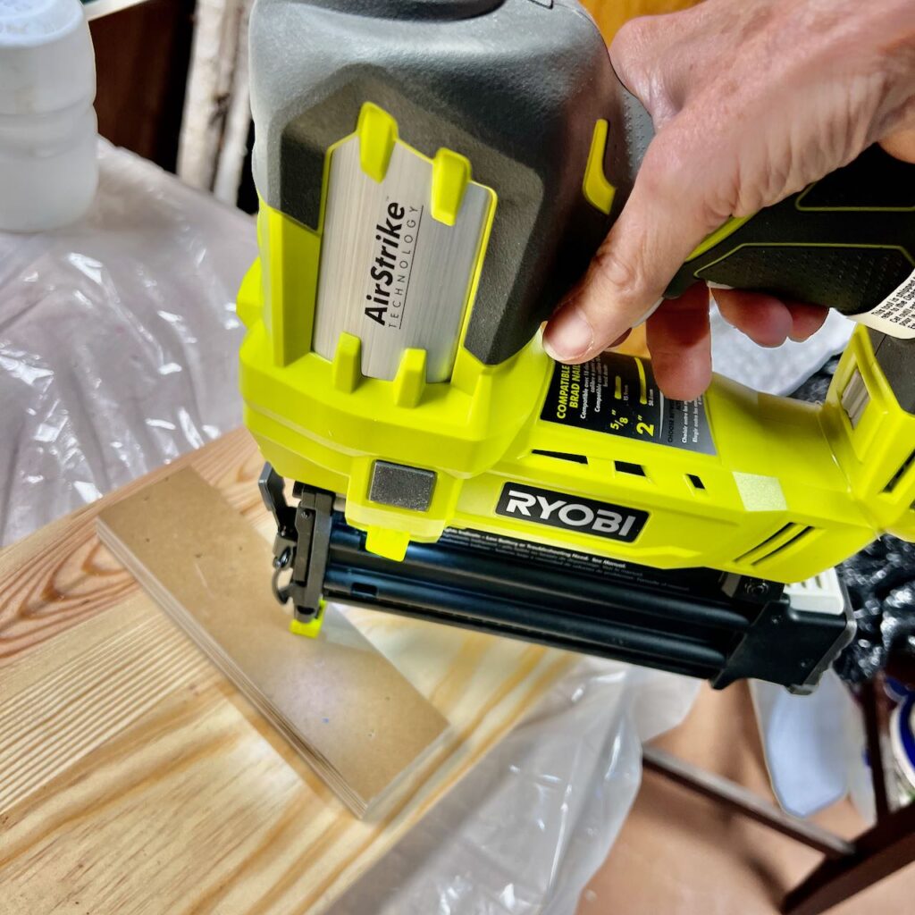 a woman's hand is attaching a scrap piece of wood across two wood boards with a brad nailer.