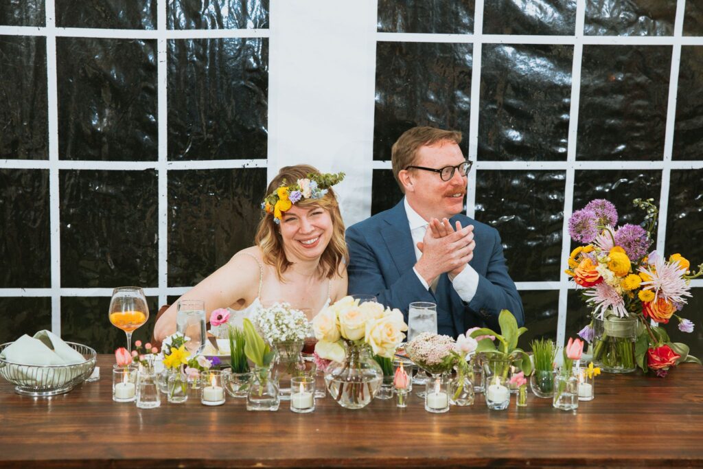 The bride and groom are seated at their wood bridal table with a long cluster centerpiece down the center of it and the bride's bouquet. off to the side