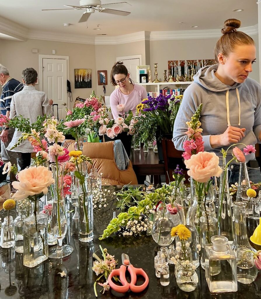 A woman standing between a table full of buckets of flowers and a counter with three cluster arrangements being assembled.The counter hassome vases with flowers and some still waiting, scissors and leaves and bits of stems