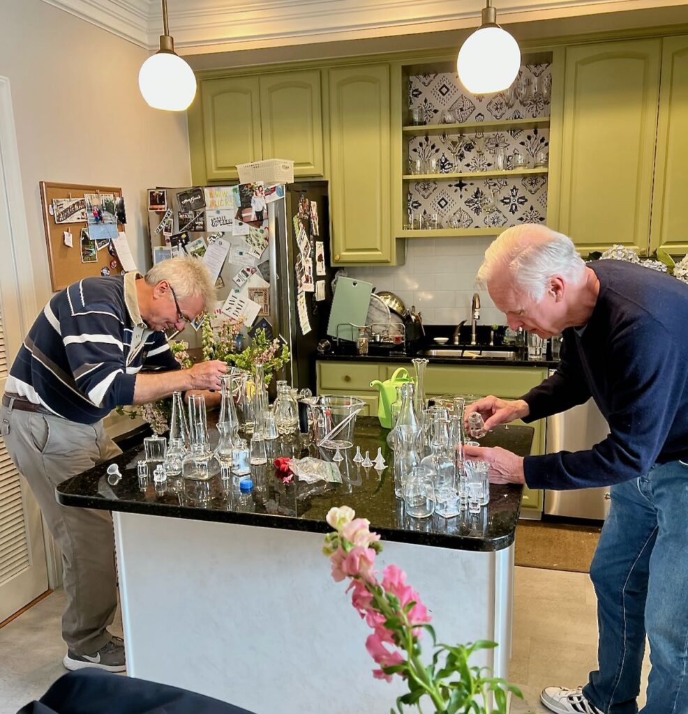 Two gentlemen shown concentrating to fill the empty tiny bud vases of three cluster centerpieces with water prior to their being filled with flowers for the
