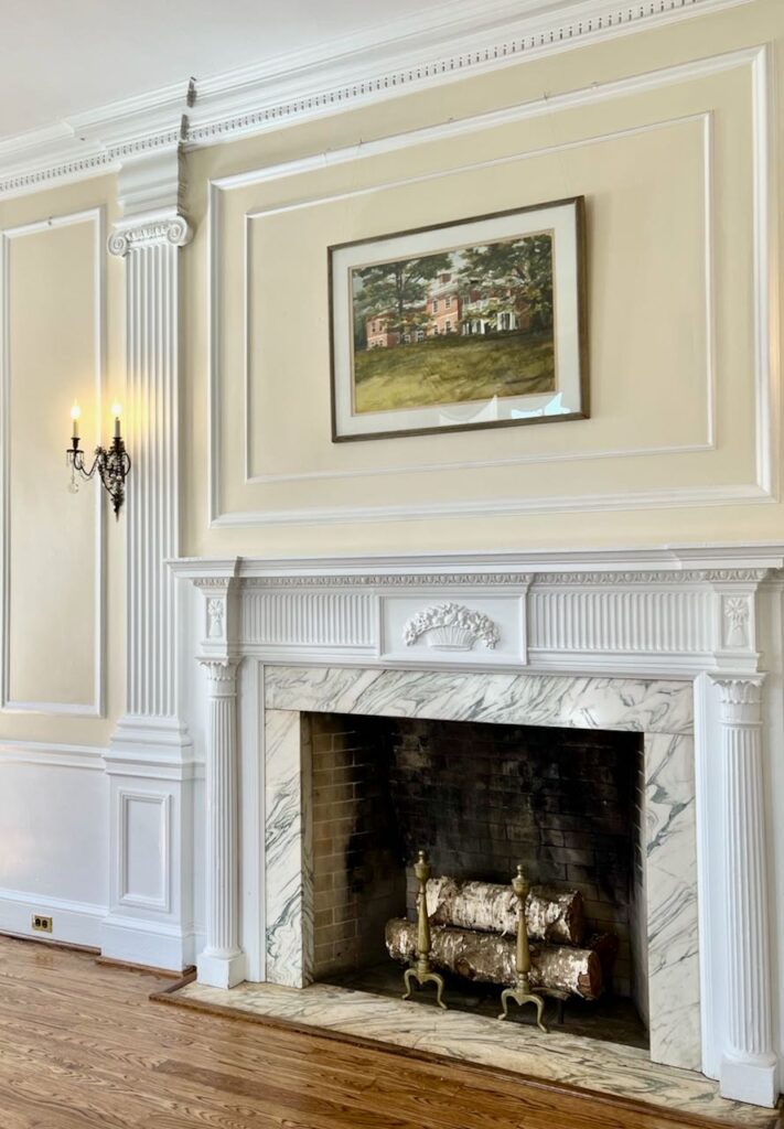 image of an ornate white fireplace mantel and surround with ornate moulding above and a framed landscape