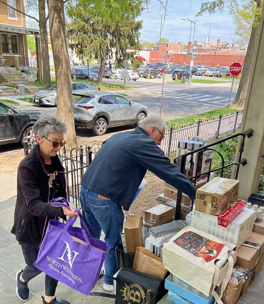 Image of an older couple delivering piles of boxes and bags onto the front porch of a large home