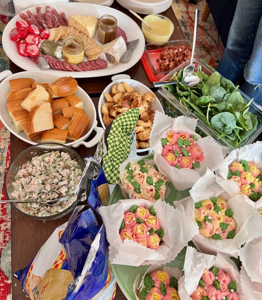 Overhead view of a food spread on a table including a meat and cheese plate, salad greens, chicken salad, small rolls and beautiful flower decorated cupcakes