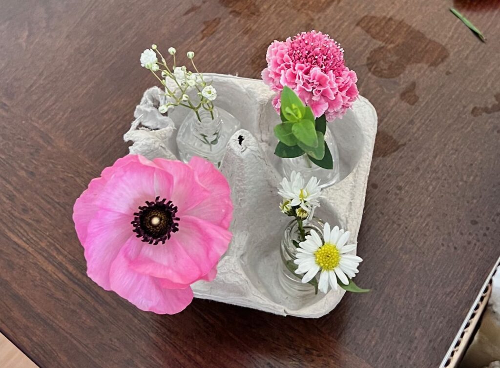 four micro bud vases are shown each with a bloom nestled in a four-part section of an egg carton