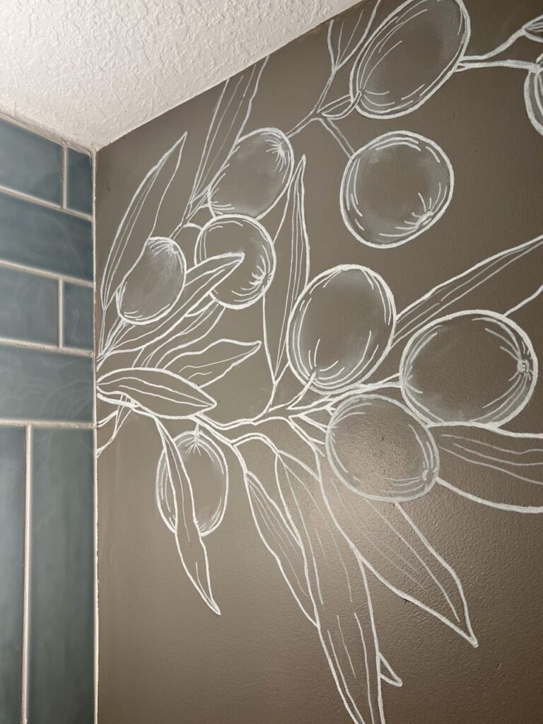 Image of an upper corner of the powder room with a blue tile wall on one side and the other wall is a dark taupe wall with a white chalk painted mural of olive branches