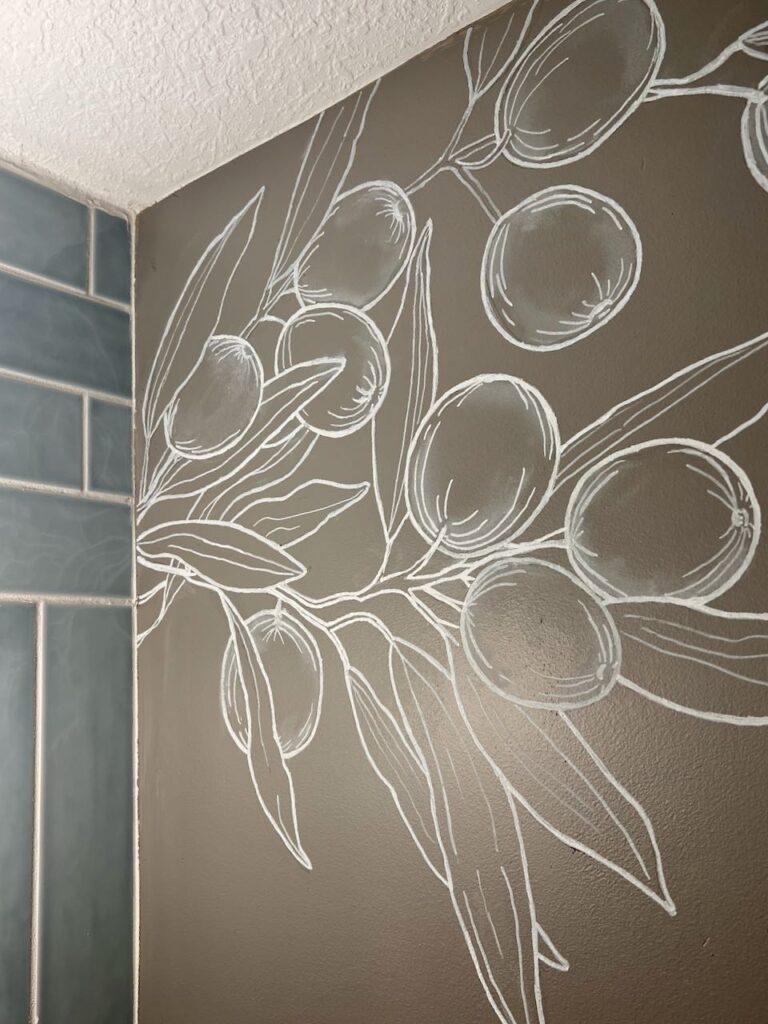 Image of an upper corner of the powder room with a blue tile wall on one side and the other wall is a dark taupe wall with a white chalk painted mural of olive branches