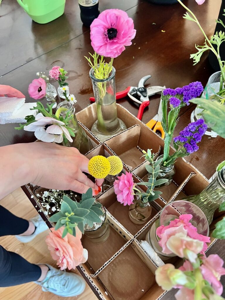 the inside of a box that has been fitted with dividers cut from corrugated cardboard partially filled with a variety of bud vases each holding a single bloom. A hand is placing another vase in the box and adding pieces of styrofoam to keep it all packed nicely.