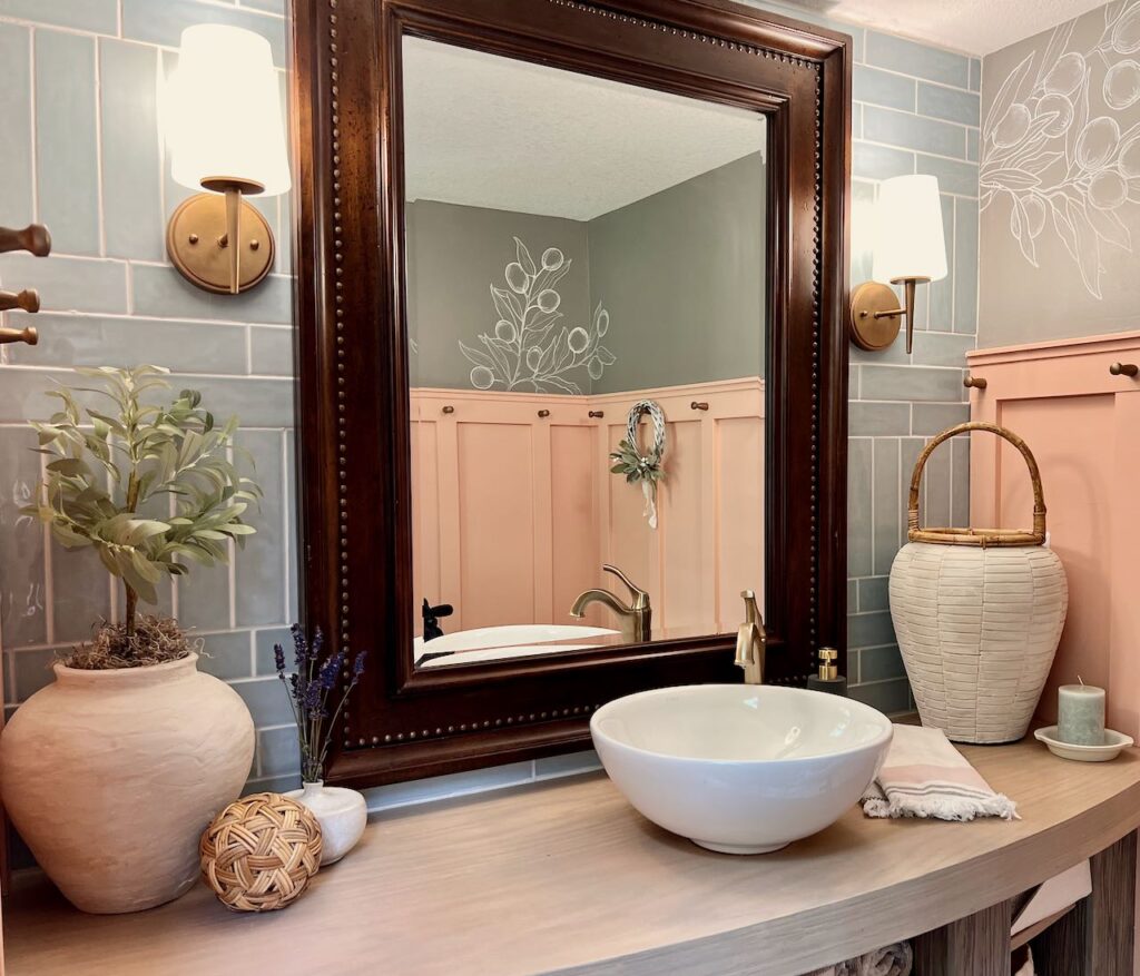 Image of the finished powder room remodel showing the vanity top with sink, faucet and decor pieces in front of a blue tile wall and a large framed mirror. Reflected in the mirror is the coral board and batten wall with gold pegs on on one peg is hanging a small wreath accented with dried lambs ear