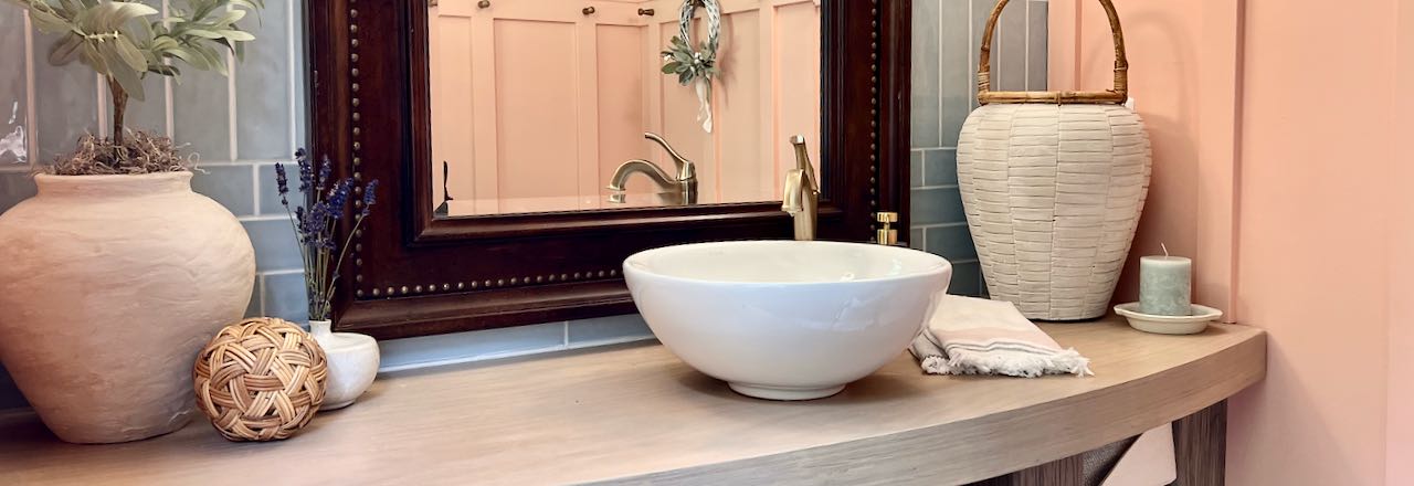 Title image of the vanity top with sink and decor pieces in front of a blue tile wall and a large framed mirror. Reflected in the mirror is the coral board and batten wall with gold pegs on on one peg is hanging a small wreath accented with dried lambs ear