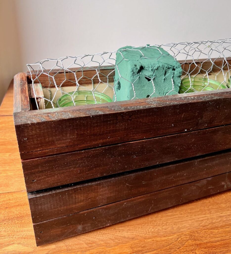 Closeup. of a third of the long wooden box filled with bell jars, florist foam and a chicken wire dome