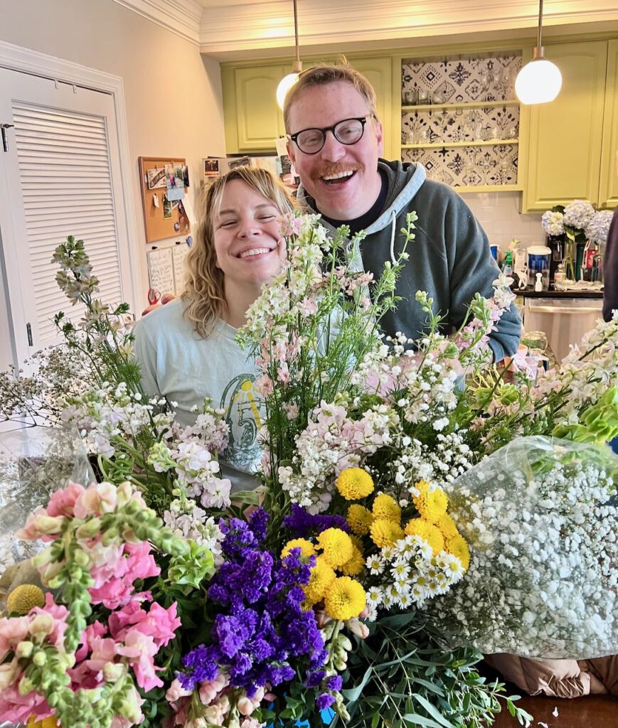 Peeking out behind a forest of flowers is a young smiling couple, our bride and groom