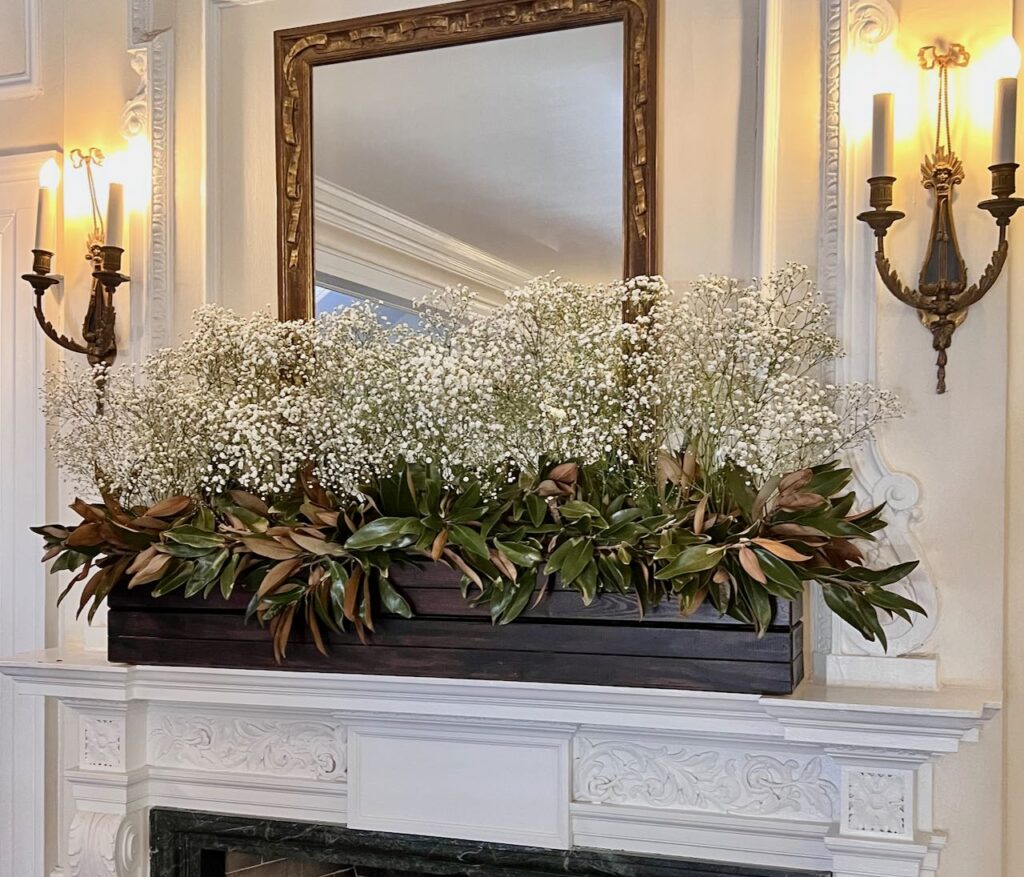 Image of a long dark wood planter box filled with magnolia leaves and Baby's Breath centered on an ornate white mantel with a framed mirror above and two brass sconces on each side