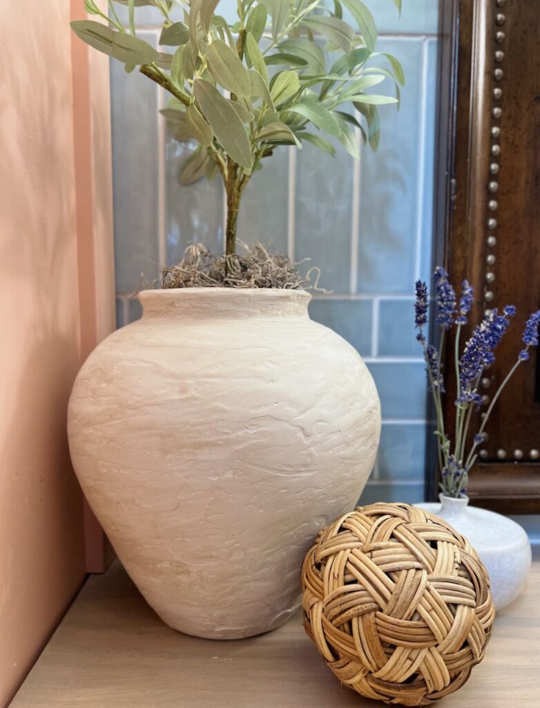 a madeover "rustic" vase holding an olive tree with a mound of spanish moss next to a rattan ball and a small vase folding dried lavender in front of a blue tile wall and a heavy wooden frame