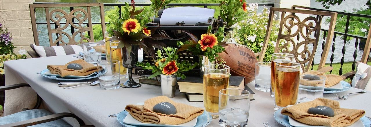 wide title shot of a Fathers Day tablescape that features a vintage typewriter, books and bottles and jiggers filled with flowers and greenery along with plates with a smooth black rock with a name written in chalk marker sitting on a folded napkin at each placesetting