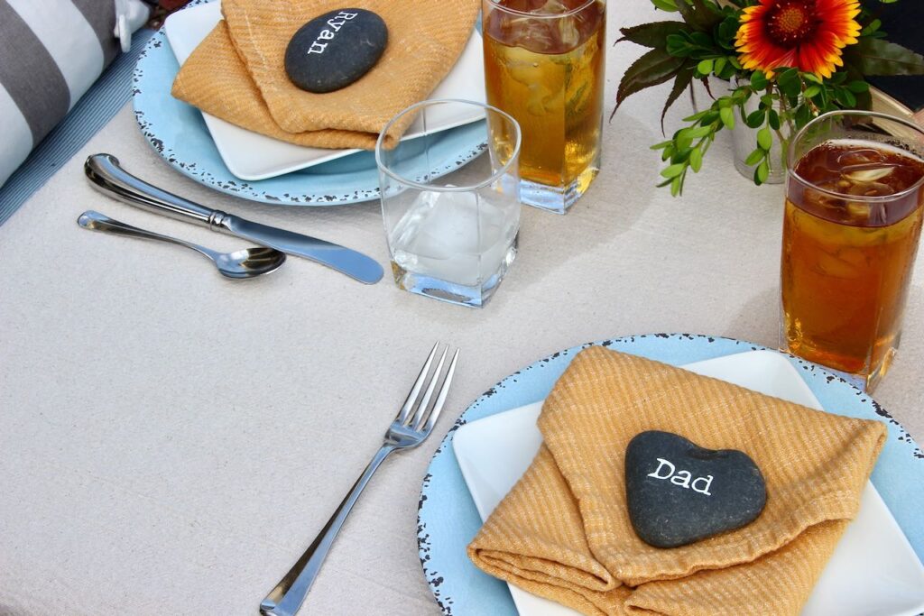 View of two placesettings on the corner of the Father's Day table. Each placesetting includes a blue dinner plate with a square white salad plate, a gold napkin folded with a smooth black rock with names written in a typewriter font on top