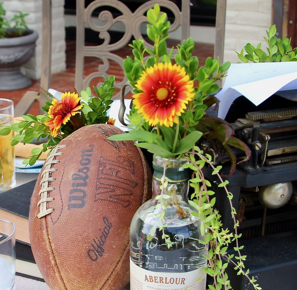 Closeup of flowers , greenery and trailing ground cover in an empty Scotch bottle next to a football