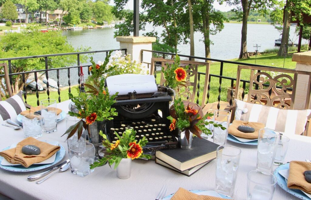 view of a table overlooking a lake, set with a long neutral tablecloth and a centeriece featuring a vintage typewriter, various books, a football, an apollo model and five bottles and jiggers filled with flowers and greenery