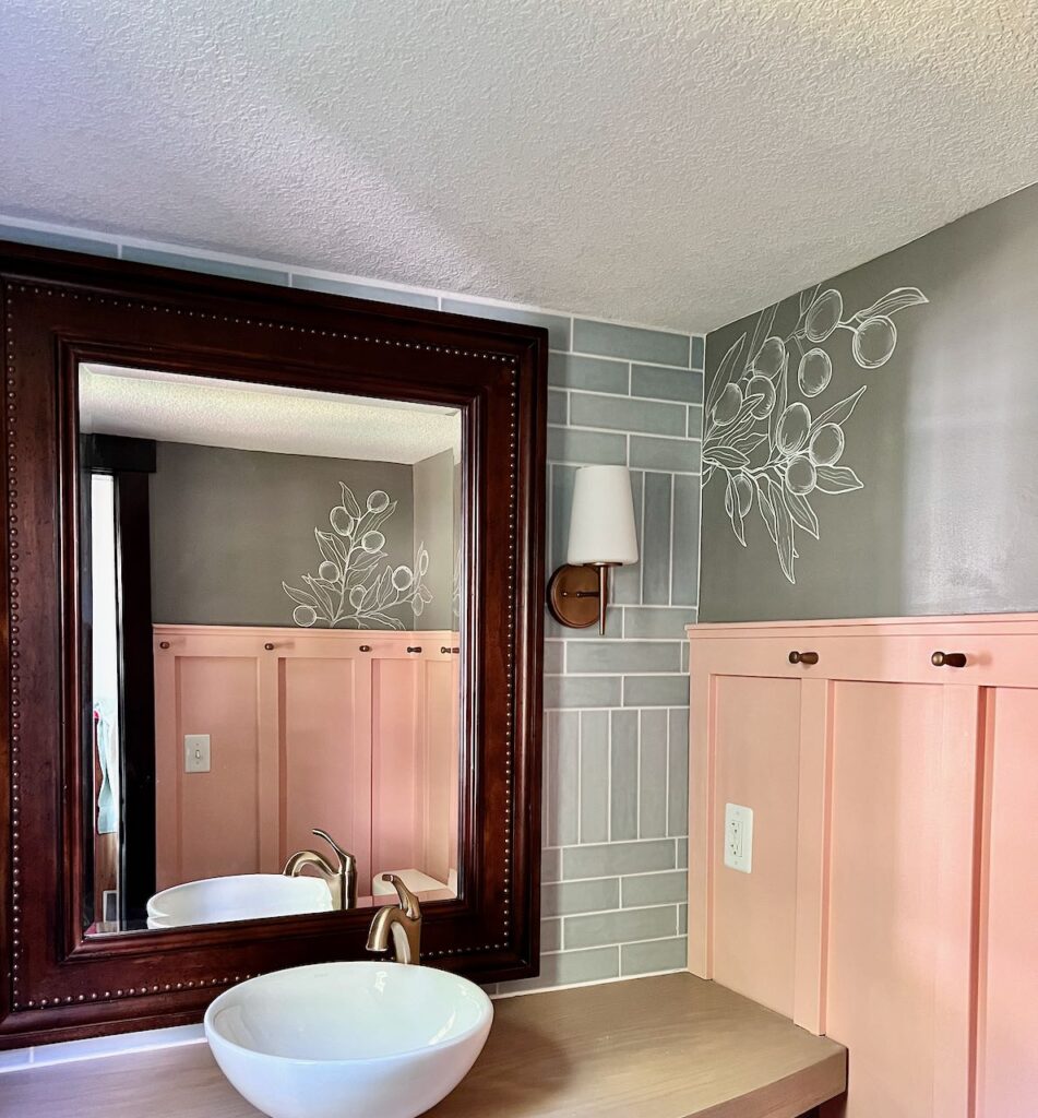 View into powder room with light coming in through the window, one cluster of olives branches is seen straight ahead to the right of the sink and another cluster is seen reflected in the mirror above the sink