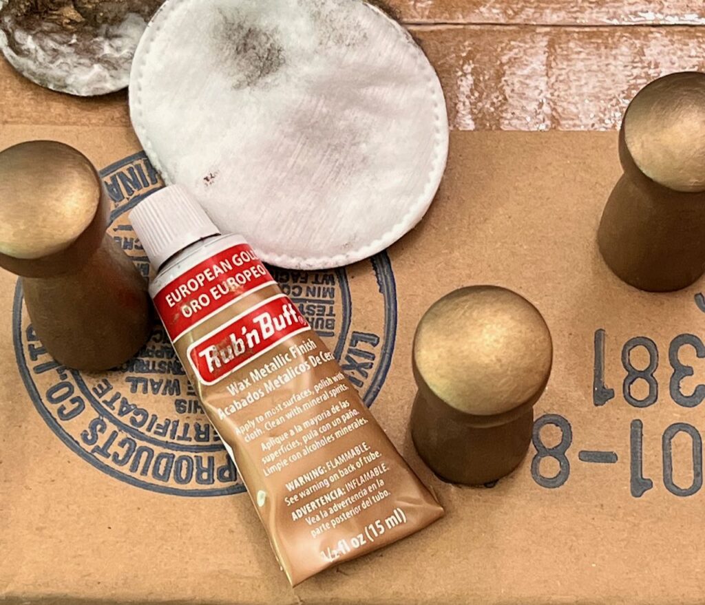 three wood pegs are shown with a tube of European Gold Rub 'n Buff wax next to them and used facial pads. The pegs are standing on a box to dry.