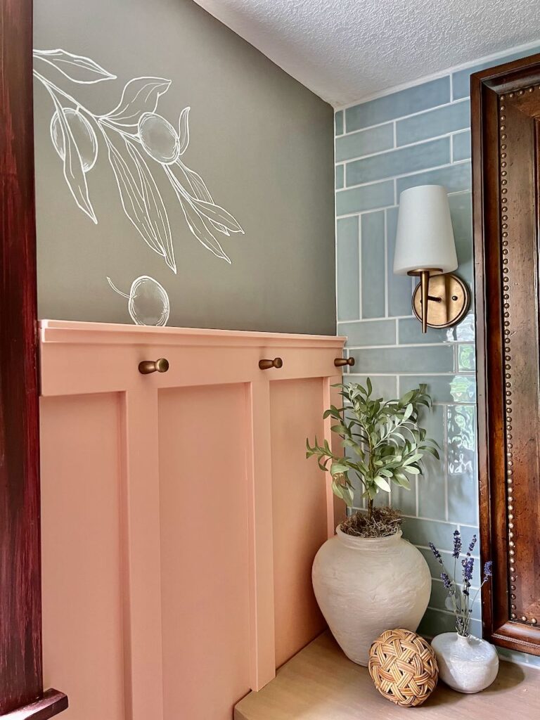 View of line art of olive branch cluster peeking out from window and an olive fallen off the branch is resting on the board and batten accent wall ledge