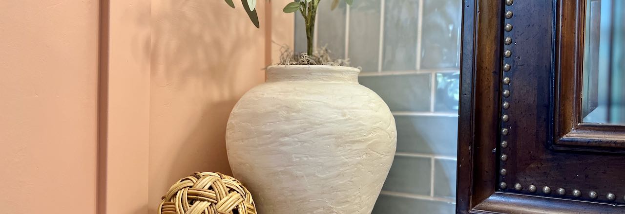 wide view. of large rustic vase dupe holding an olive tree next to a rattan ball and a small vase with dried lavender in front of a blue tile wall and a heavy wood framed mirror