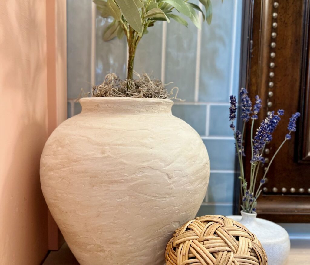 view. of large rustic vase dupe holding an olive tree next to a rattan ball and a small vase with dried lavender in front of a blue tile wall and a heavy wood framed mirror