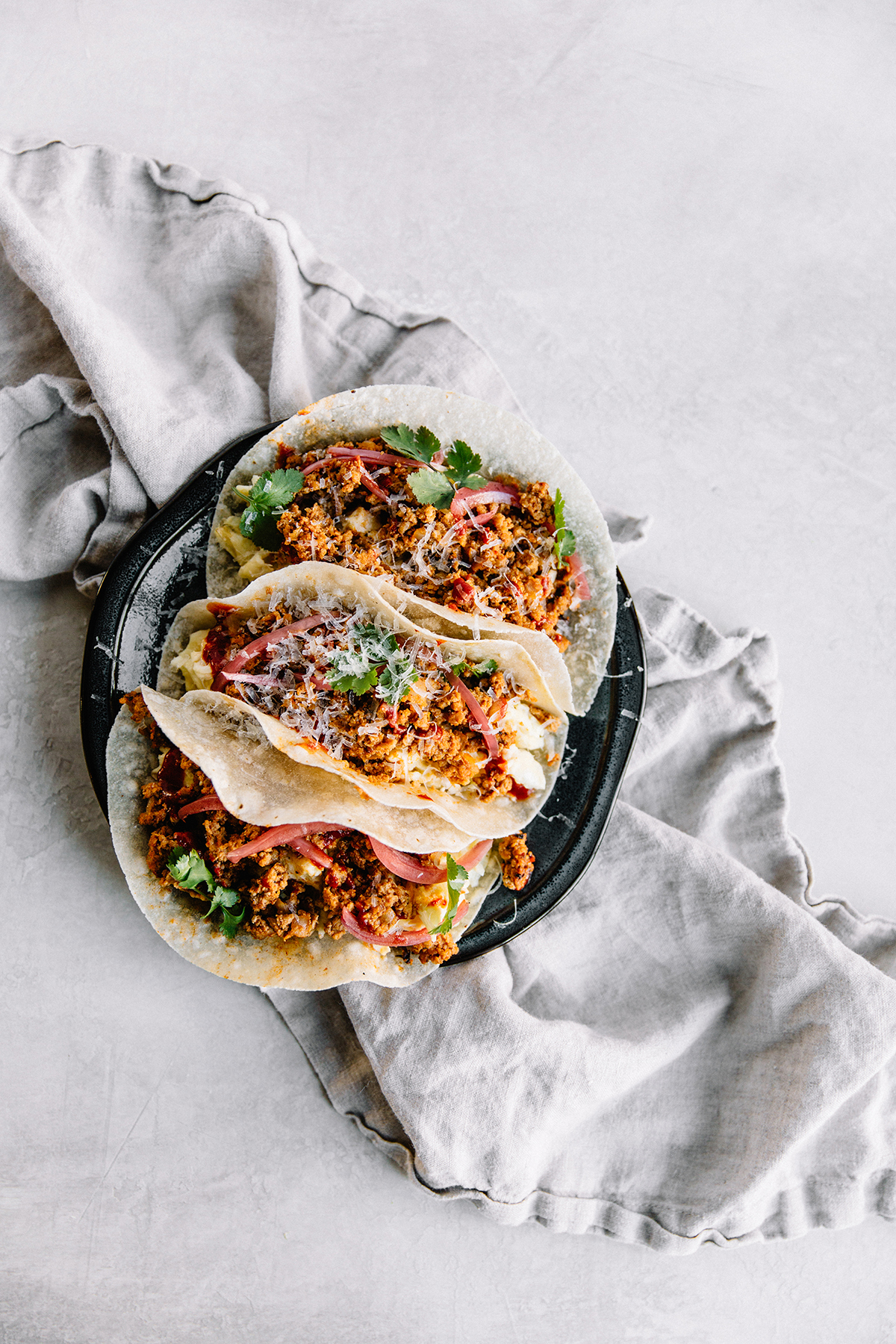 Three soft tacos filled with seasoned ground meat, topped with fresh cilantro, onions, and grated cheese, served on a black plate over a light gray cloth on a light-colored background.