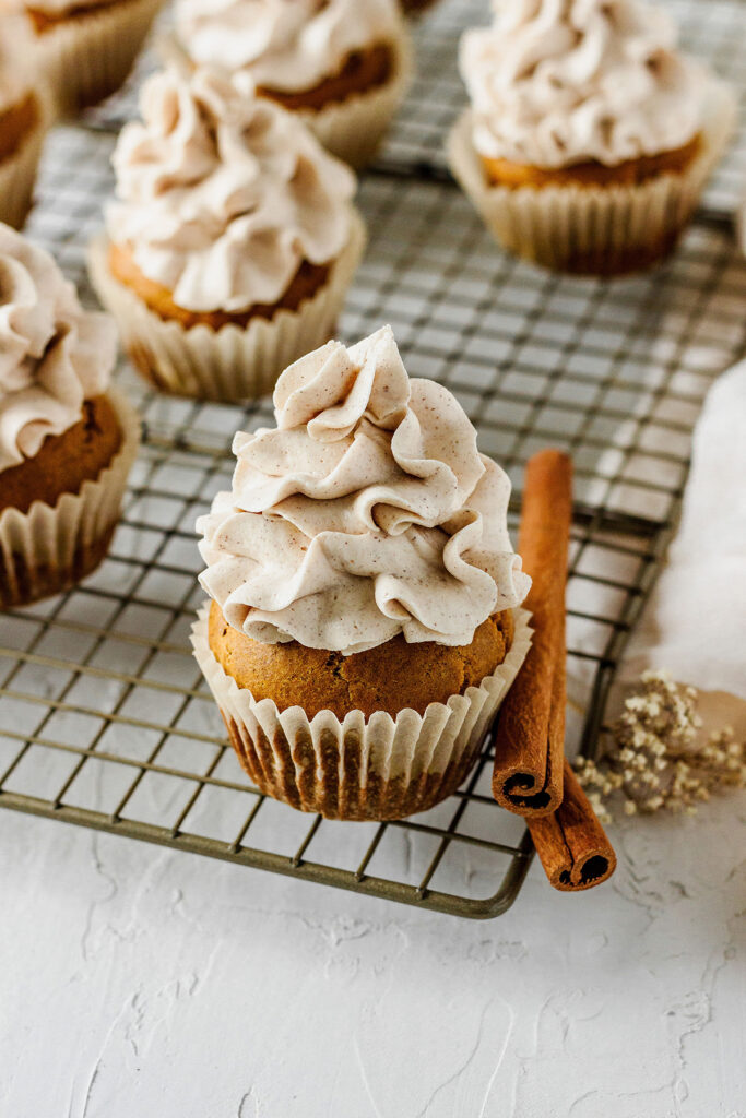 A cupcake topped with a generous swirl of cinnamon frosting sits on a cooling rack beside two cinnamon sticks, with more cupcakes in the background.