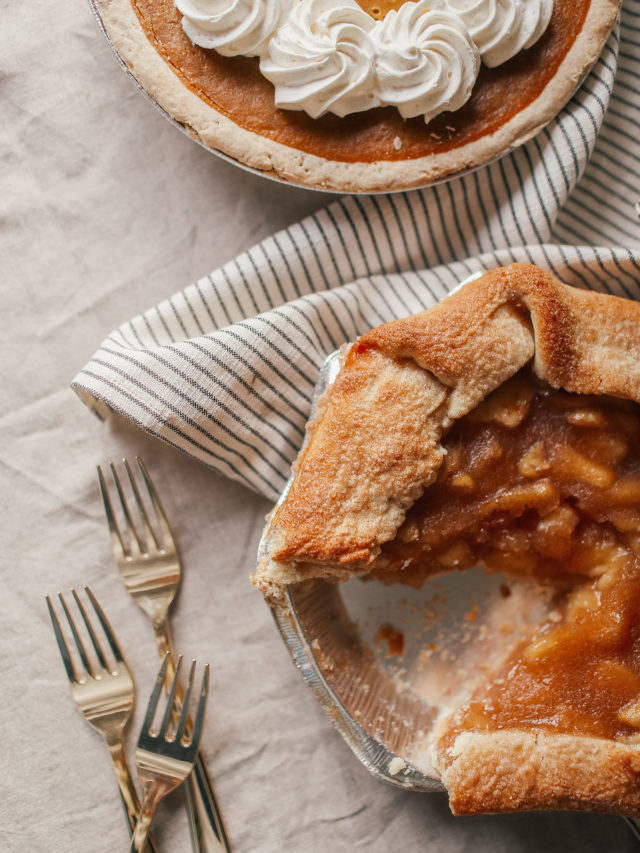 Overhead view of a partially eaten apple pie in a foil pan and a whole pie with whipped cream topping, placed on a striped cloth with three forks nearby.