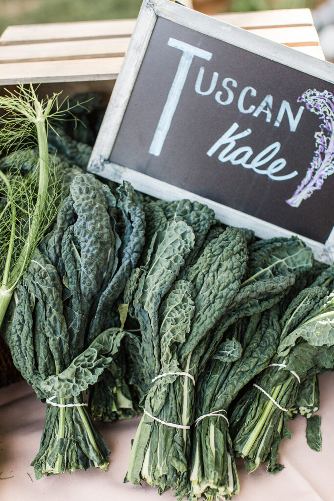 Bunches of Tuscan kale are displayed on a table, tied with string, next to a rustic chalkboard sign labeled "Tuscan kale" with a hand-drawn kale illustration.