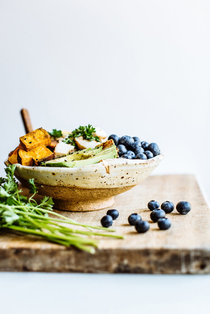 A ceramic bowl on a wooden board, filled with roasted sweet potatoes, sliced avocado, grilled chicken, blueberries, and parsley, with some blueberries and parsley scattered nearby.